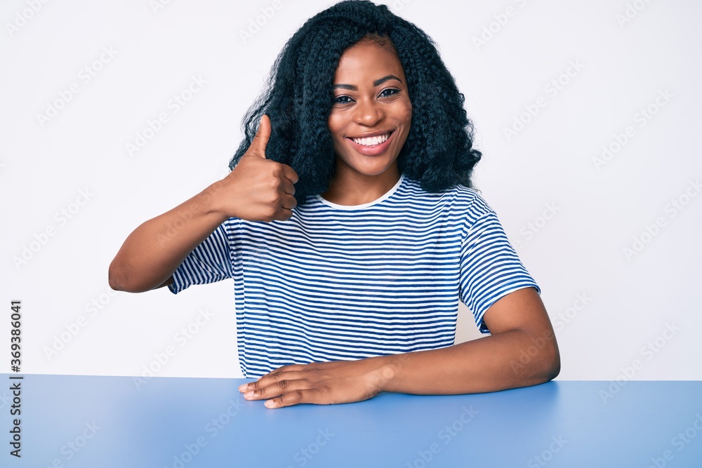 Beautiful african woman wearing casual clothes sitting on the table smiling happy and positive, thumb up doing excellent and approval sign Beautiful african woman wearing casual clothes sitting on the table smiling happy and positive, thumb up doing excellent and approval sign