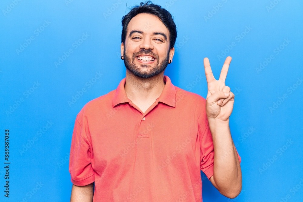 Fototapeta premium Young hispanic man wearing casual clothes smiling with happy face winking at the camera doing victory sign. number two.