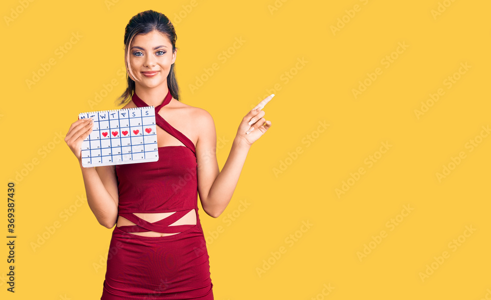 Young beautiful woman holding heart calendar smiling happy pointing with hand and finger to the side Young beautiful woman holding heart calendar smiling happy pointing with hand and finger to the side
