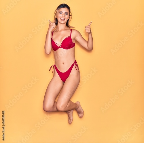 Young beautiful girl on vacation wearing bikini smiling happy. Jumping with smile on face doing ok sign with thumbs up over isolated yellow background