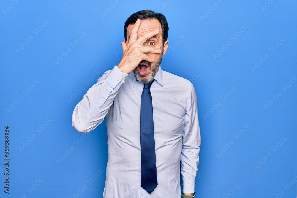 Middle age handsome business man wearing elegant tie standing over isolated blue background peeking in shock covering face and eyes with hand, looking through fingers afraid Middle age handsome business man wearing elegant tie standing over isolated blue background peeking in shock covering face and eyes with hand, looking through fingers afraid