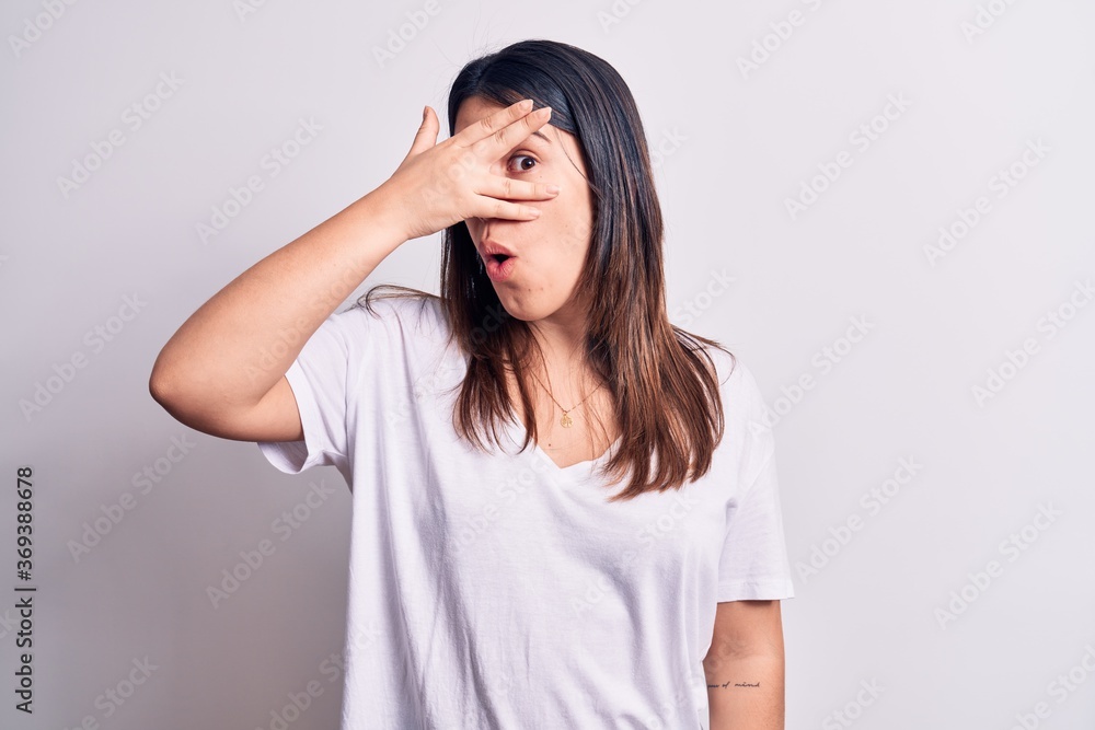 Young beautiful brunette woman wearing casual t-shirt standing over isolated white background peeking in shock covering face and eyes with hand, looking through fingers afraid Young beautiful brunette woman wearing casual t-shirt standing over isolated white background peeking in shock covering face and eyes with hand, looking through fingers afraid