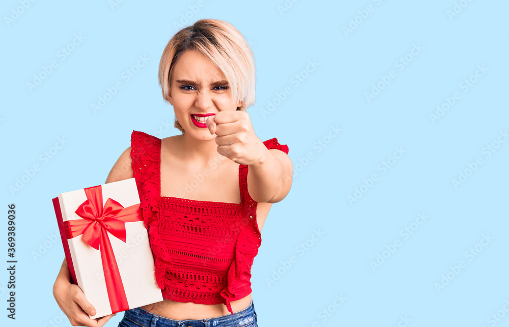 Young beautiful blonde woman holding gift annoyed and frustrated shouting with anger, yelling crazy with anger and hand raised Young beautiful blonde woman holding gift annoyed and frustrated shouting with anger, yelling crazy with anger and hand raised