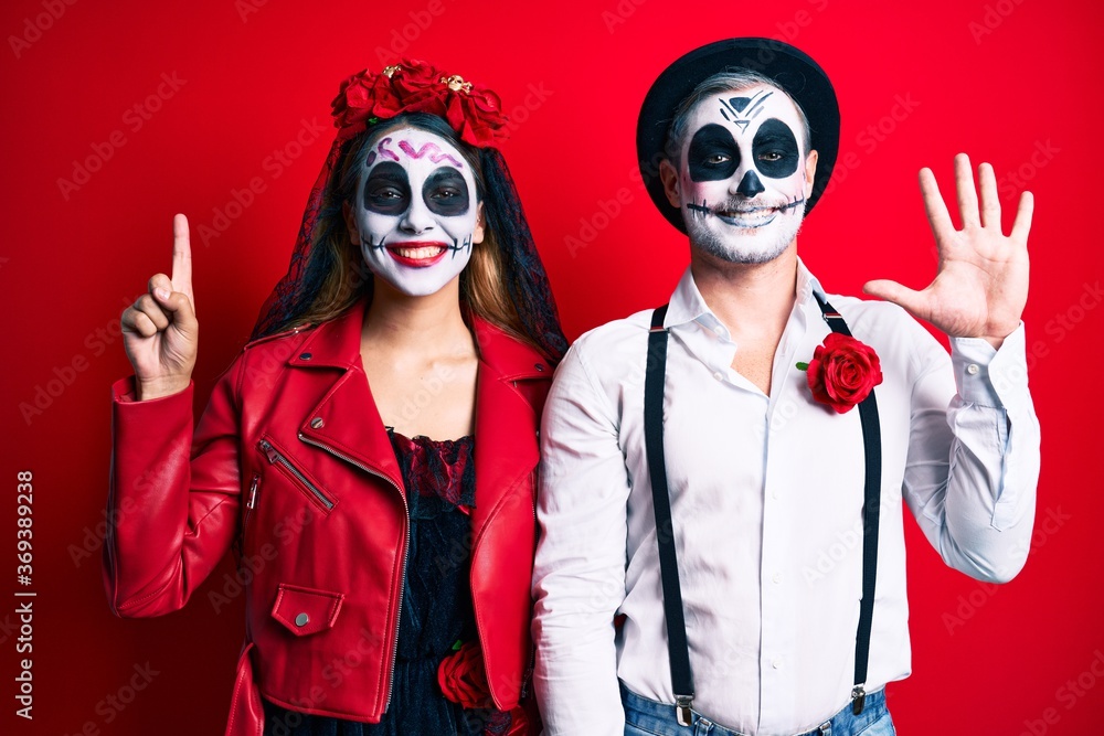 Couple wearing day of the dead costume over red showing and pointing up with fingers number six while smiling confident and happy.
