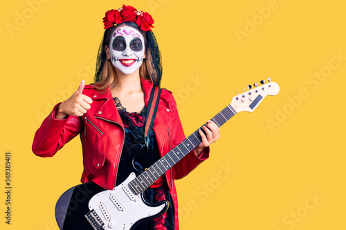 Woman wearing day of the dead costume playing electric guitar smiling happy and positive, thumb up doing excellent and approval sign