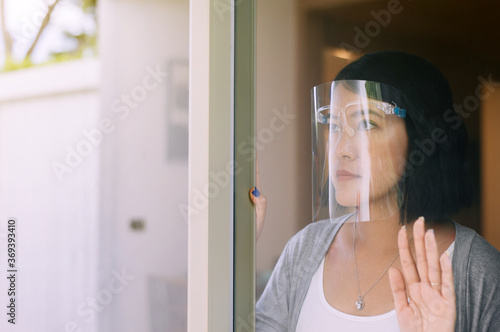 Asian woman wearing face shield with hands touching on window,Stay at home during coronavirus and covid-19 epidemic or pandemic,Social distancing concept