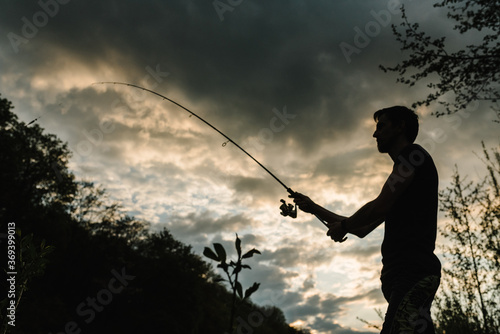 Silhouette of a fisherman. Young man fishing on a lake at sunset. Fishery, fishing day. Rod rings, fishing tackle. Fisherman with rod, spinning reel on the river bank. Fishing for pike, perch, carp.