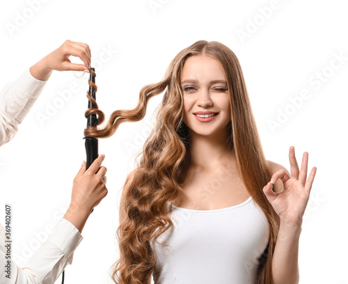 Hairdresser curling hair of beautiful young woman against white background