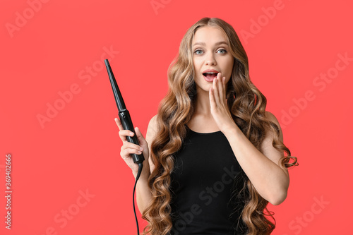Vászonkép Surprised young woman with curling iron for hair against color background
