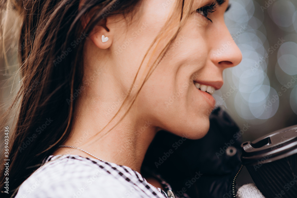 Fototapeta premium A young beautiful woman in mittens smiles and holds a disposable cup of coffee in her hands against the background of Christmas lights garlands on a windy winter day. Soft selective focus, art noise
