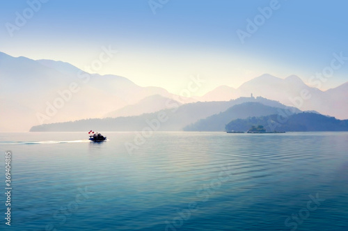 Scenic landscape, view over blue lake and small tourist boat, travelling along calm water, layers of mountains over the background in the tranquil morning, Sun Moon lake, Taiwan