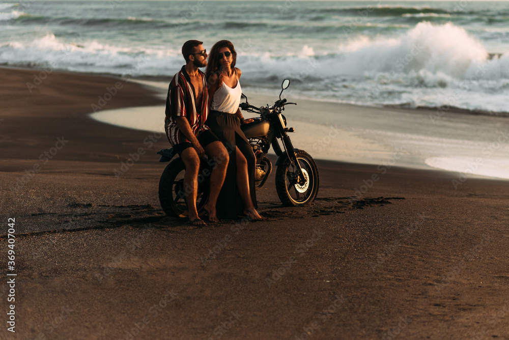 Obraz premium Zakochana para na plaży spotyka zachód słońca. Piękna para na motocyklu spotyka zachód słońca nad morzem. Mężczyzna i kobieta podróżują na rowerze po Bali. Mężczyzna i kobieta o zachodzie słońca. Wakacje na morzu.