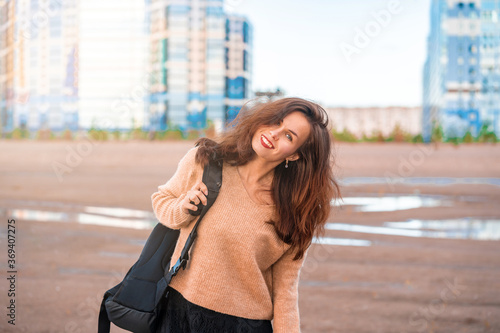 A smiling young woman in a cozy sweater and skirt with a backpack walks on a sandy wasteland with a view of multi-storey new buildings, dreams of buying their own housing