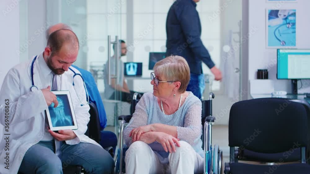 Doctor holding tablet pc with x-ray while explaining diagnosis to disabled elderly woman in wheelchair. Handicapped patient in hospital waiting area. Man in examination room. Doctor holding tablet pc with x-ray while explaining diagnosis to disabled elderly woman in wheelchair. Handicapped patient in hospital waiting area. Man in examination room.