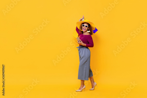 Full length portrait of happy young attractive African American woman raising shopping bags and holding mobile phone in isolated studio yellow background