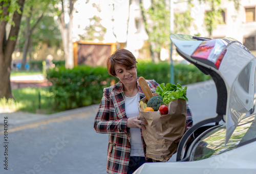 Beautiful middle age woman taking out from car trunk paper bag full of fresh vegetables and fruit, smiling. Carrying a healthy bag. 