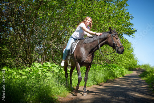 A fat girl and a brown horse in a Park on a Sunny day and green trees in the background. Young woman plus size rides a horse.