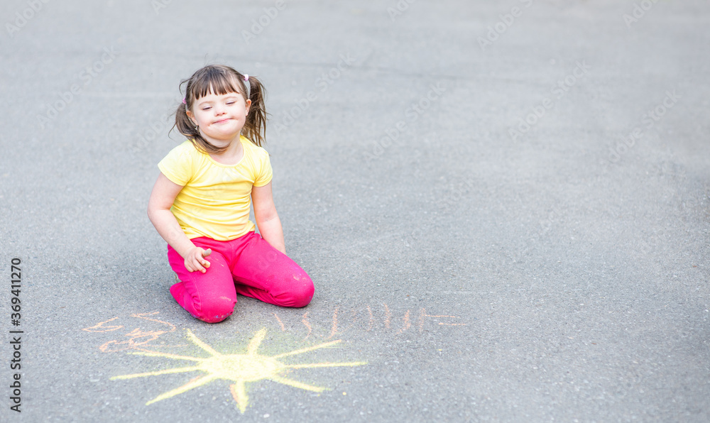 Smiling girl with syndrome down draws the sun with chalk on the asphalt. Empty space for text Smiling girl with syndrome down draws the sun with chalk on the asphalt. Empty space for text