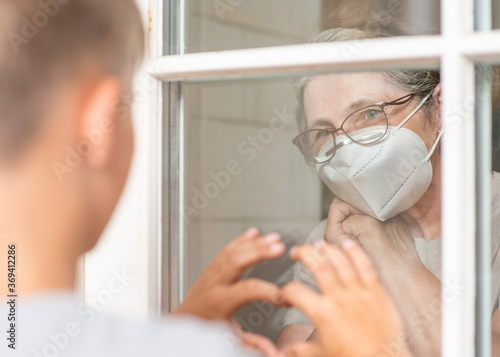 Grandmother wearing protective mask communicates with her grandson through a window during an epidemic of coronavirus. Grandson shows heart sign to grandmother