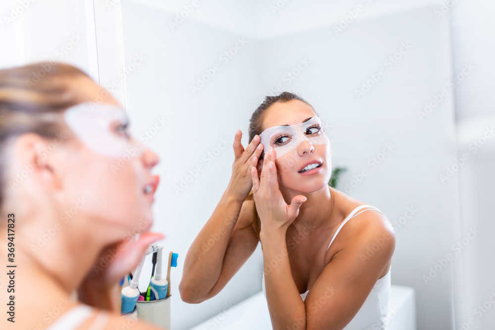 Woman is applying sheet mask on her face in the bathroom. Skin care girl touch patches of fabric mask under eyes to reduce eye bags. Woman is applying sheet mask on her face in the bathroom. Skin care girl touch patches of fabric mask under eyes to reduce eye bags.