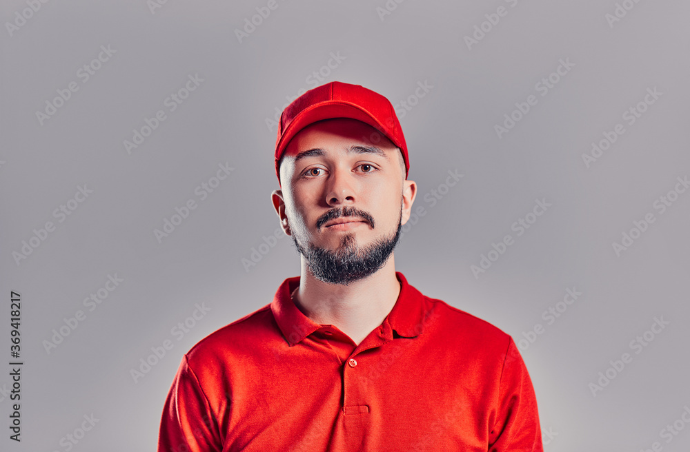 Bearded young delivery man in red t-shirt and cap isolated on gray background. Home delivery service. Bearded young delivery man in red t-shirt and cap isolated on gray background. Home delivery service.