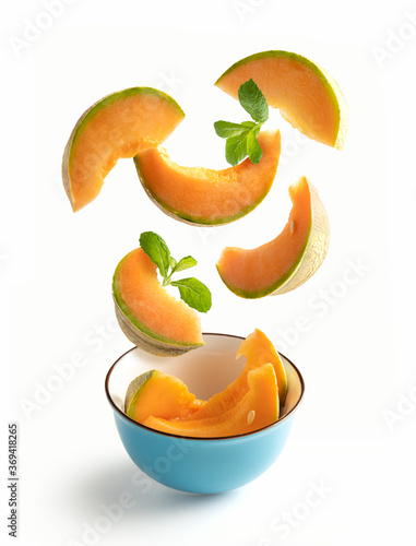 Cantaloupe melon slices with flying mint leaves in a blue bowl, isolated from white background