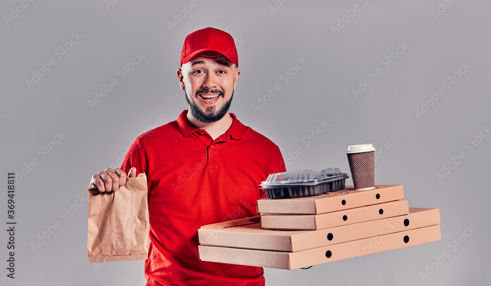 Bearded young delivery man in red T-shirt and cap with pizza boxes and paper fast food bag and cups of coffee isolated on gray background. Fast home delivery. Bearded young delivery man in red T-shirt and cap with pizza boxes and paper fast food bag and cups of coffee isolated on gray background. Fast home delivery.