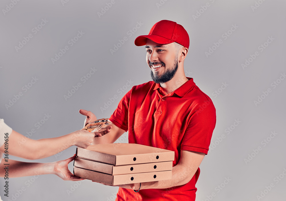 Young bearded delivery man in red uniform delivers pizza to a client and takes money isolated on gray background. Young bearded delivery man in red uniform delivers pizza to a client and takes money isolated on gray background.