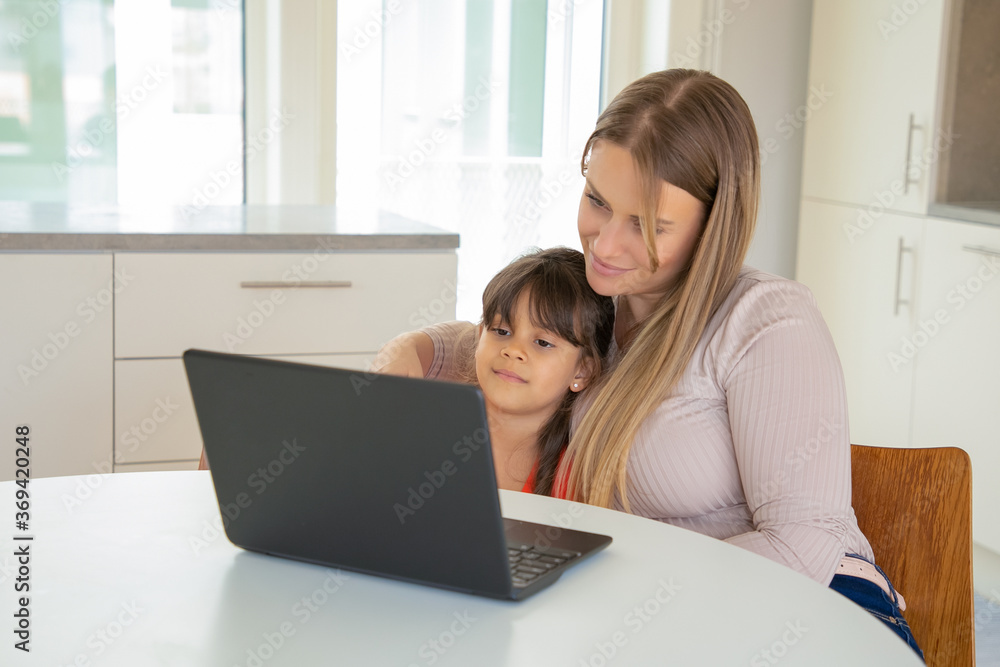 Peaceful girl and her mom using laptop, sitting at table and hugging, watching movie, looking at display. Front view. Internet and communication concept Peaceful girl and her mom using laptop, sitting at table and hugging, watching movie, looking at display. Front view. Internet and communication concept