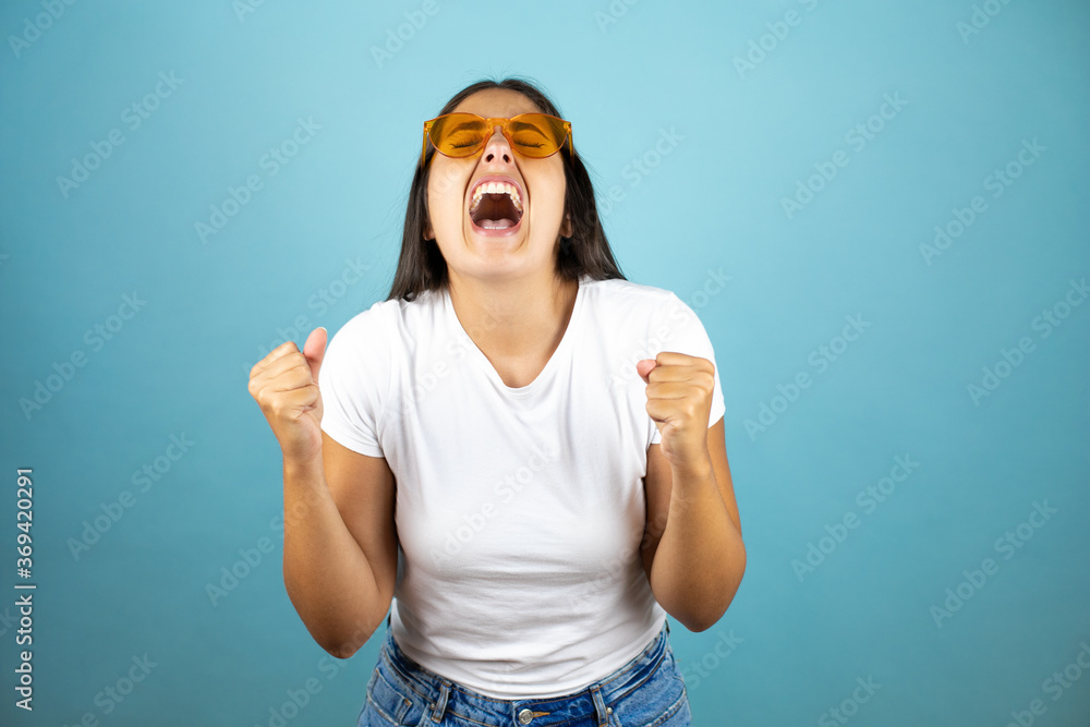 Young beautiful woman wearing sunglasses standing over isolated blue background screaming with her fists in the air with a gesture of victory Young beautiful woman wearing sunglasses standing over isolated blue background screaming with her fists in the air with a gesture of victory