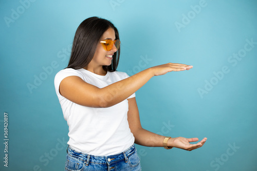 Young beautiful woman wearing sunglasses over isolated blue background smiling and gesturing with hands showing big and large size sign, measure symbol