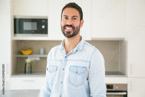 Cheerful attractive dark haired Latin man posing in kitchen, looking at camera and smiling. Medium shot, copy space. Man at home or male portrait concept