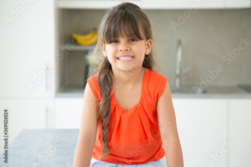 Cheerful dark haired Latin little girl wearing red sleeveless shirt, posing in kitchen, looking at camera and smiling. Medium shot, front view. Childhood or child portrait concept