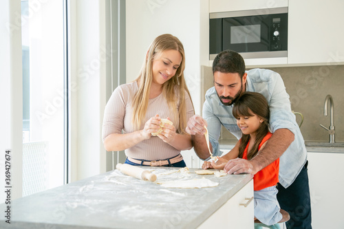 Happy mom and dad teaching daughter to knead dough at kitchen table with flour powder. Young couple and their girl baking together. Family cooking concept