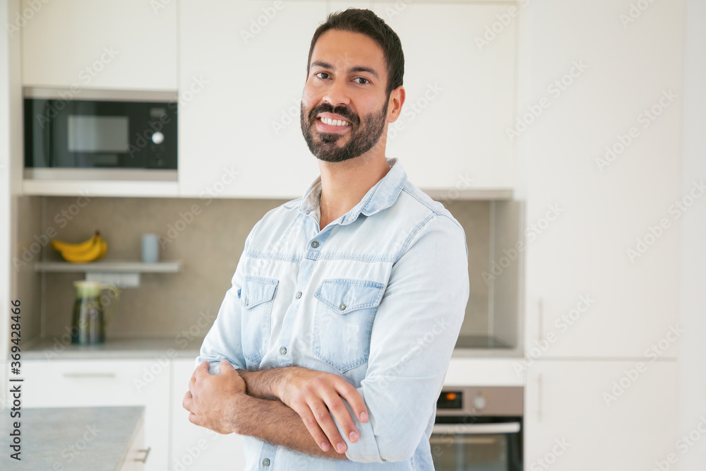 Happy handsome dark haired Latin man posing with arms folded in kitchen, looking at camera and smiling. Medium shot, copy space. Man at home or male portrait concept