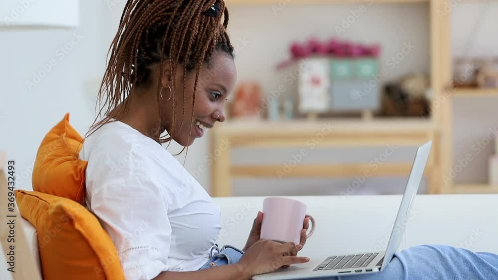 Young happy woman is using laptop while sitting on sofa in apartment interior spbi. Closeup view of African American female looking at computer screen with smile and browsing web on couch in light Young happy woman is using laptop while sitting on sofa in apartment interior spbi. Closeup view of African American female looking at computer screen with smile and browsing web on couch in light