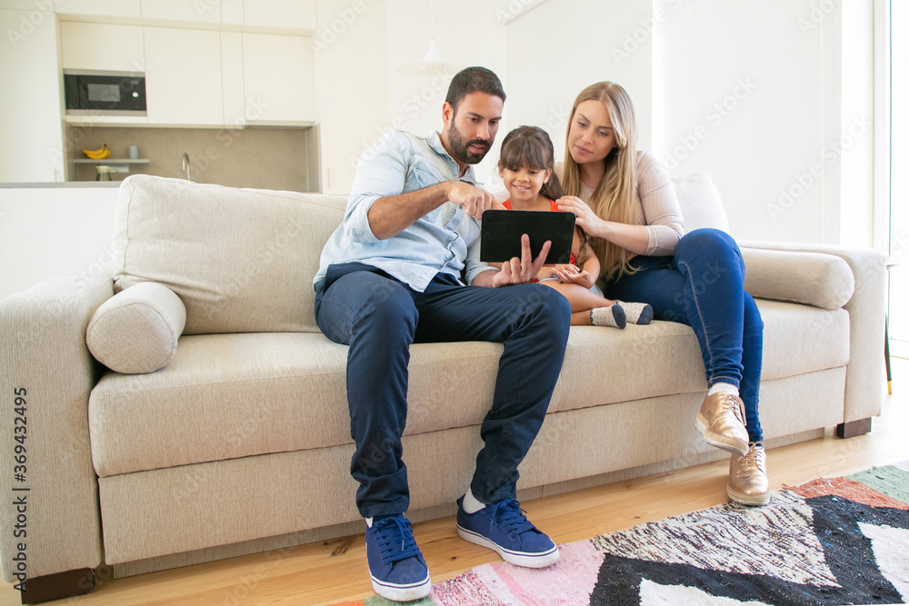 Happy family sitting on couch, using online app on tablet, looking at screen, watching movie together. Internet and communication concept