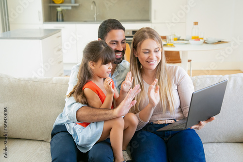 Happy parents and cute daughter using laptop for video call, talking to grandparents, waving hello, looking at display while sitting on couch at home. Communication during pandemic concept