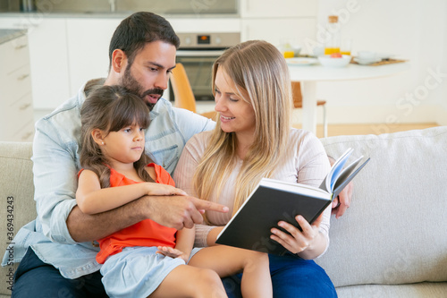 Happy parents couple and little black haired girl sitting on couch in living room and reading book together. Childhood or literature concept