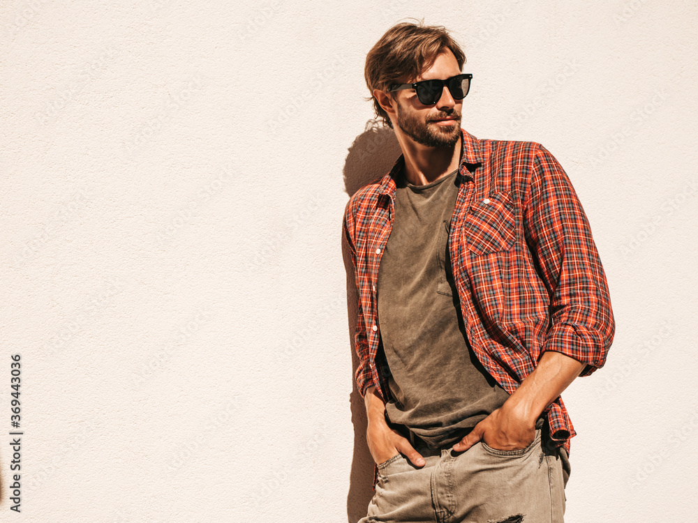 Portrait of handsome stylish hipster lambersexual model.Man dressed in checkered shirt. Fashion male posing in the street near white wall outdoors in sunglasses