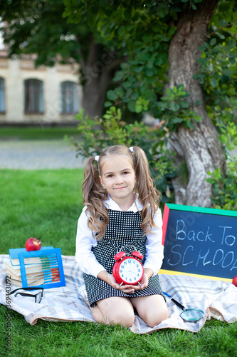 Back to school. Smiling caucasian schoolgirl with big red alarm clock relax on lawn in park near school. Kid with alarm clock in hand. Child and time. Education Concept. Cute girl holding Clock.  © stock_studio