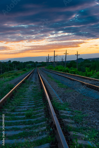 Railroad against the backdrop of sky and sunset clouds with green grass in the foreground. Beautiful sunset with orange sunbeams on a background of blue clouds. Leaving road concept.