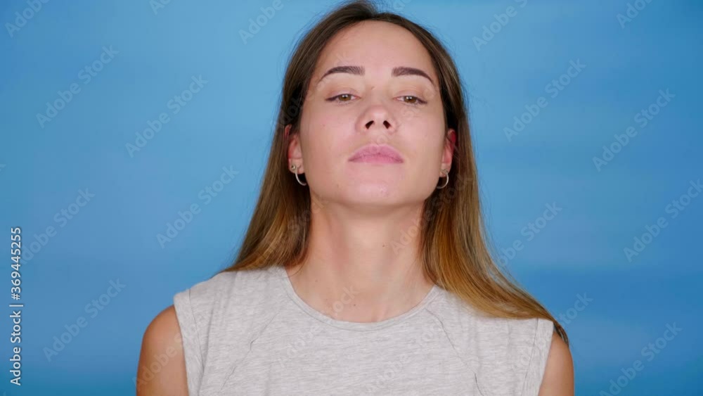 Beautiful woman in gray T-shirt scratches near the eye on blue background with copy space. 4K slow motion footage Beautiful woman in gray T-shirt scratches near the eye on blue background with copy space. 4K slow motion footage