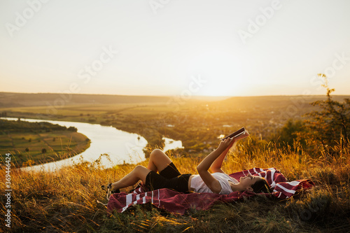 Young woman lies on red plaid on top of the mountain and reads book on sunset with perfect landscape. Copy space.