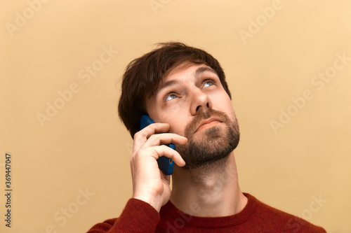 Communications. Photo of young man with beard has phone conversation, holds mobile phone near ear, tells news to friend, focused away, wears sweater, posing against yellow background photo