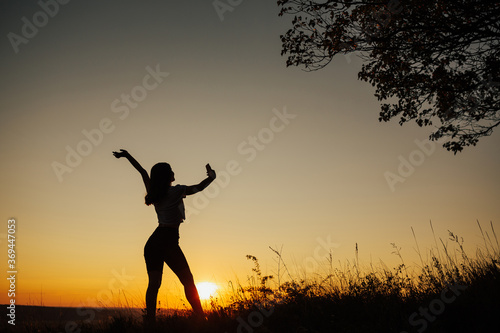 Silhouette of a woman holding a phone and taking selfie outside during sunset. Black silhouette over sunset. Woman posing on field and taking a selfie. Copy space.