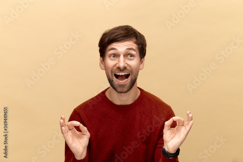 Everyting will be okay. Photo of young man with beard wearing sweater, smiling, raises okay signs, makes desirable wish, posing against yellow background. photo