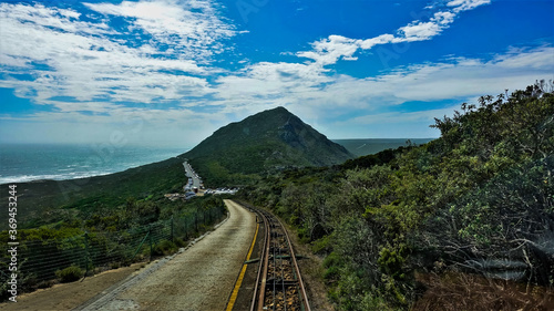 Roads in the mountains for cars and a funicular are close by. Green plants on the roadside. The highway is hidden behind a hill. The ocean is visible, the sky is bright.South Africa. Cape Point.