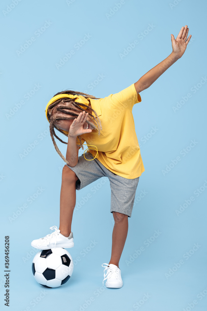 Cheerful little boy with african with headphones dabbing with one leg on soccer ball over blue background.