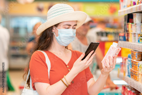 A young woman in a medical mask scans the qr code on the product using her smartphone. In the background is a supermarket. The concept of modern technology and shopping during the pandemic